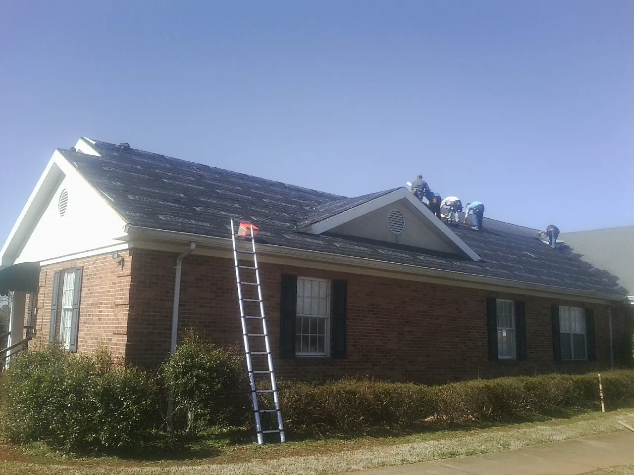 Crew tearing shingles off a brick ranch roof with a ladder set — Emergency Roof Repair work by our crew in Hacksneck