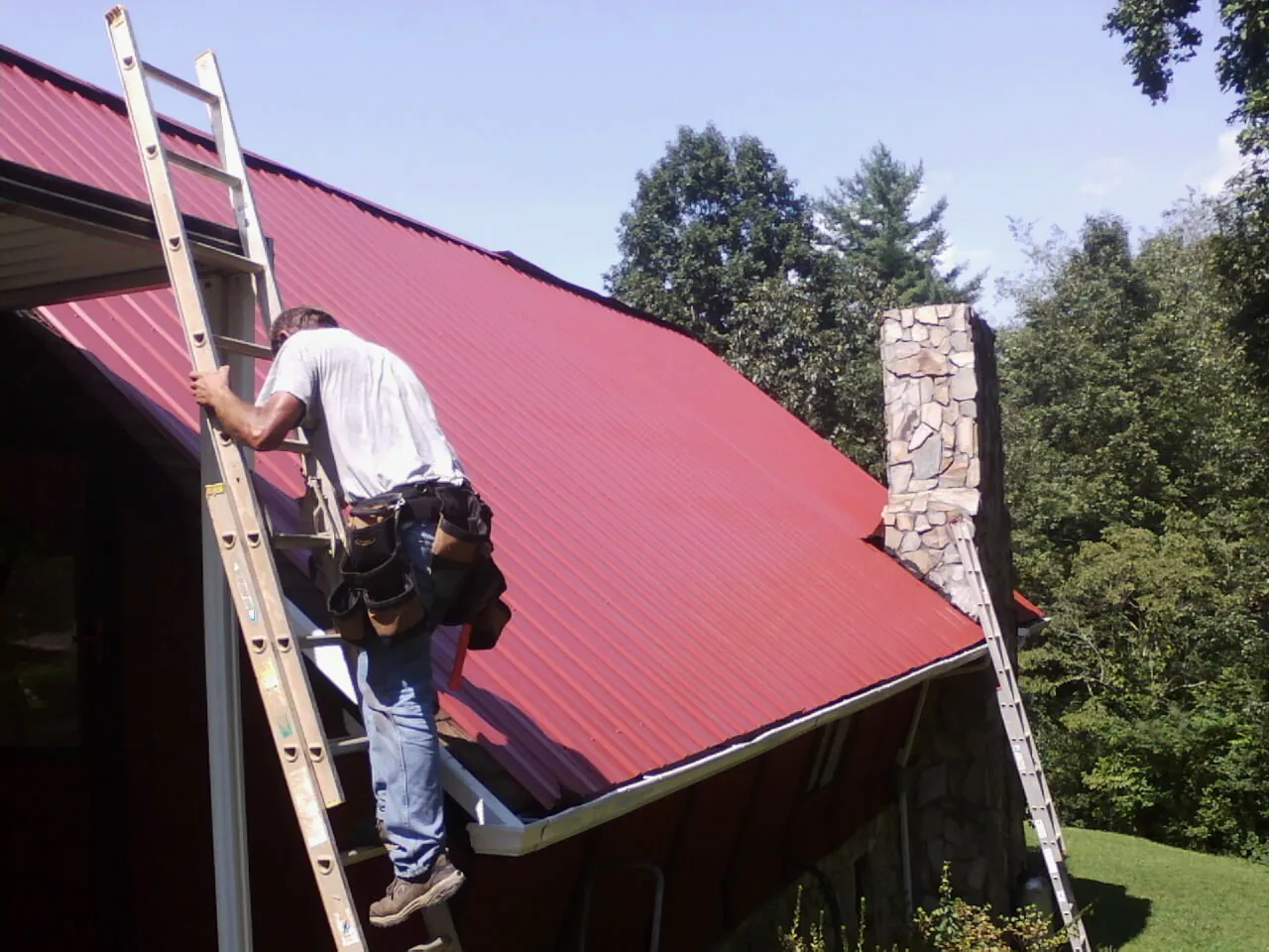 Roofer on a ladder beside a red metal roof and stone chimney — Soffit & Fascia Repair by Roofing Brothers in Hacksneck
